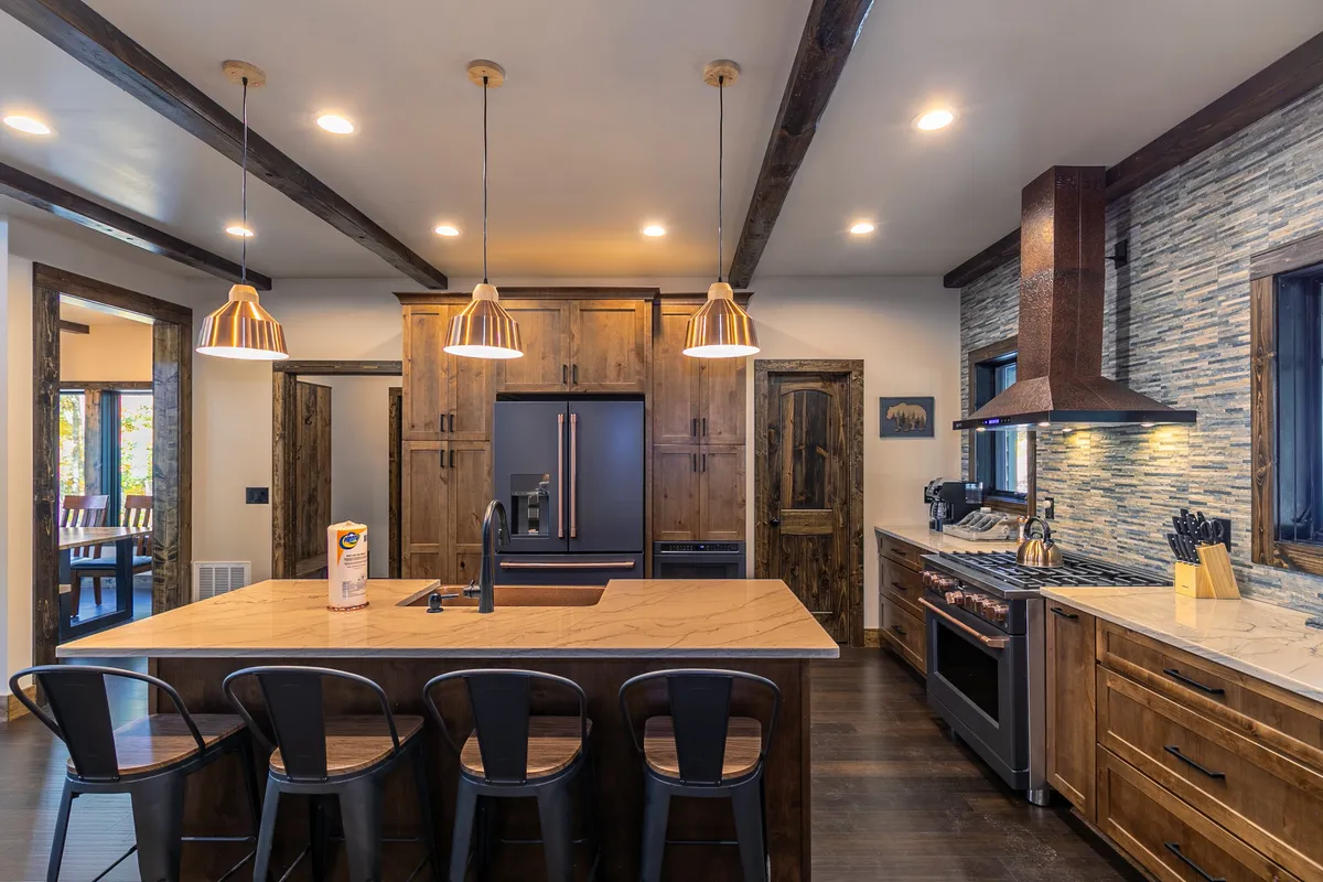 The kitchen is highlighted with a large island featuring bar seating, copper pendant lights, a stone accent wall, and stainless steel appliances. The scene feels warm and social, emphasizing the kitchen’s role as a gathering hub.