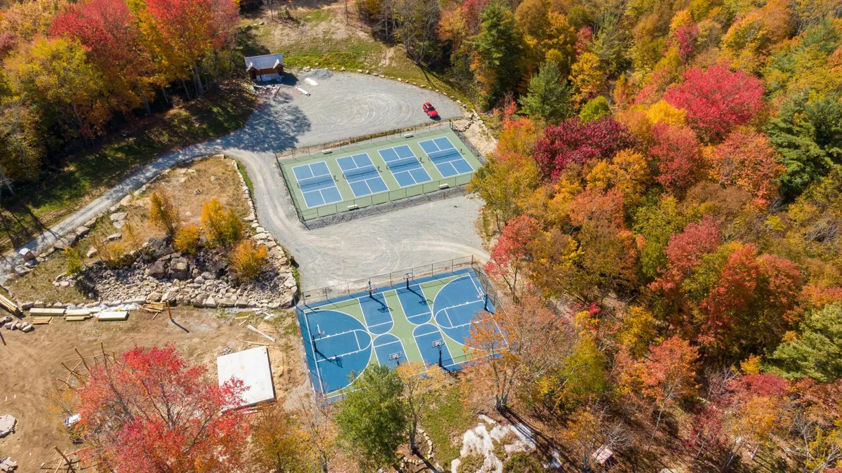 A vibrant community sports area featuring tennis and basketball courts, surrounded by colorful fall foliage and a gravel parking area. The setting is energetic and recreational, inviting guests to engage in various sports and outdoor activities.