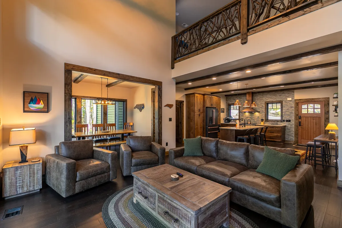 The photo reveals a wide-angle view of a cozy living room featuring plush sofas, a coffee table, a connected dining area, and a modern kitchen in the background. The space is warmly lit by stylish fixtures and infused with rustic charm, making it feel both inviting and luxurious.