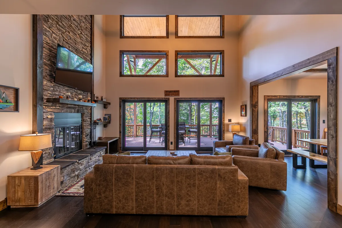 This image showcases a spacious open-concept living area with a stone fireplace, large windows streaming in natural light, comfortable seating, a television, and a dining area visible in the background. The view through the windows emphasizes the connection to the surrounding mountains and provides a luminous, airy atmosphere.