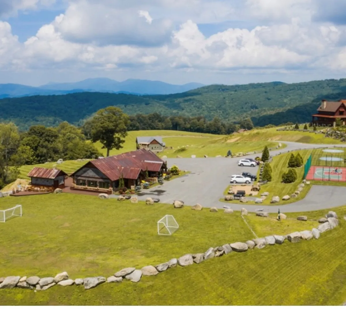 A rustic lodge sits against a mountain backdrop, with a sprawling green lawn, sports facilities, and natural rock formations creating an inviting space for families. The scene is alive with possibilities for outdoor sports, barbecues, family gatherings, and nature walks, all bathed in the glow of sunset.