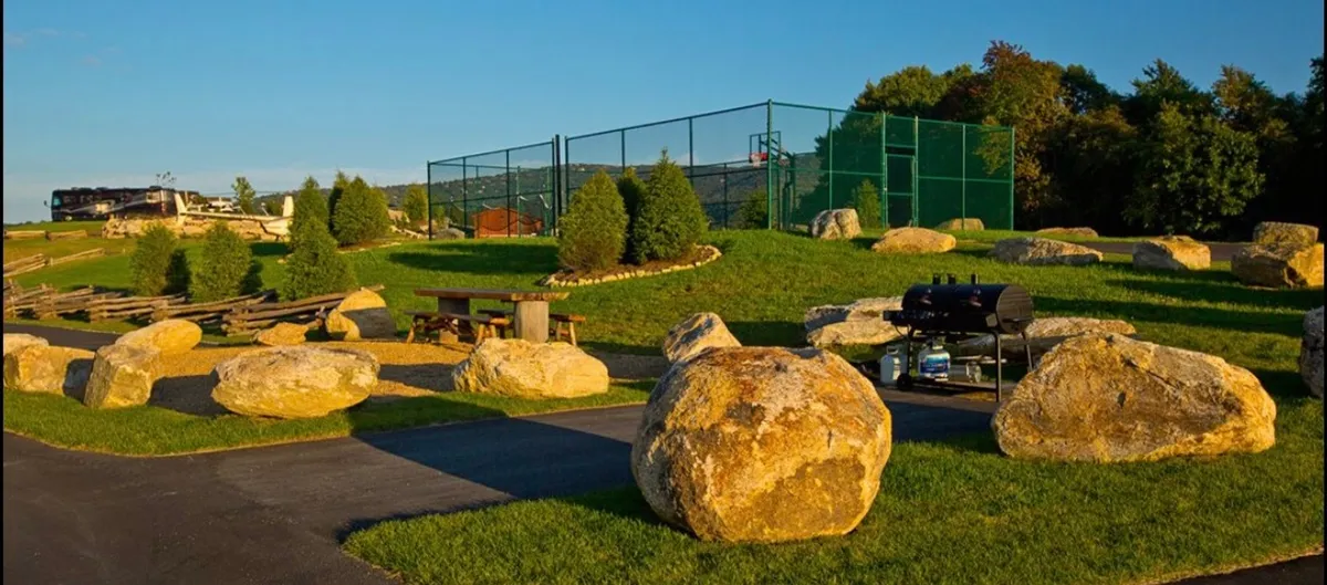 A lively outdoor amenities area with large boulders, a tennis court, a picnic area, and a grill. The space is surrounded by trees, giving it a recreational and communal vibe perfect for group activities and sports.