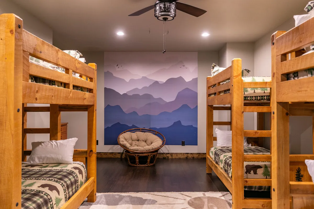 A playful bunk room designed for kids, featuring sturdy wooden bunk beds, an engaging mountain mural, a hanging ceiling fan, and a cozy round chair for reading or relaxing. The decor fosters creativity, bonding, and imaginative play – an ideal space for younger guests.