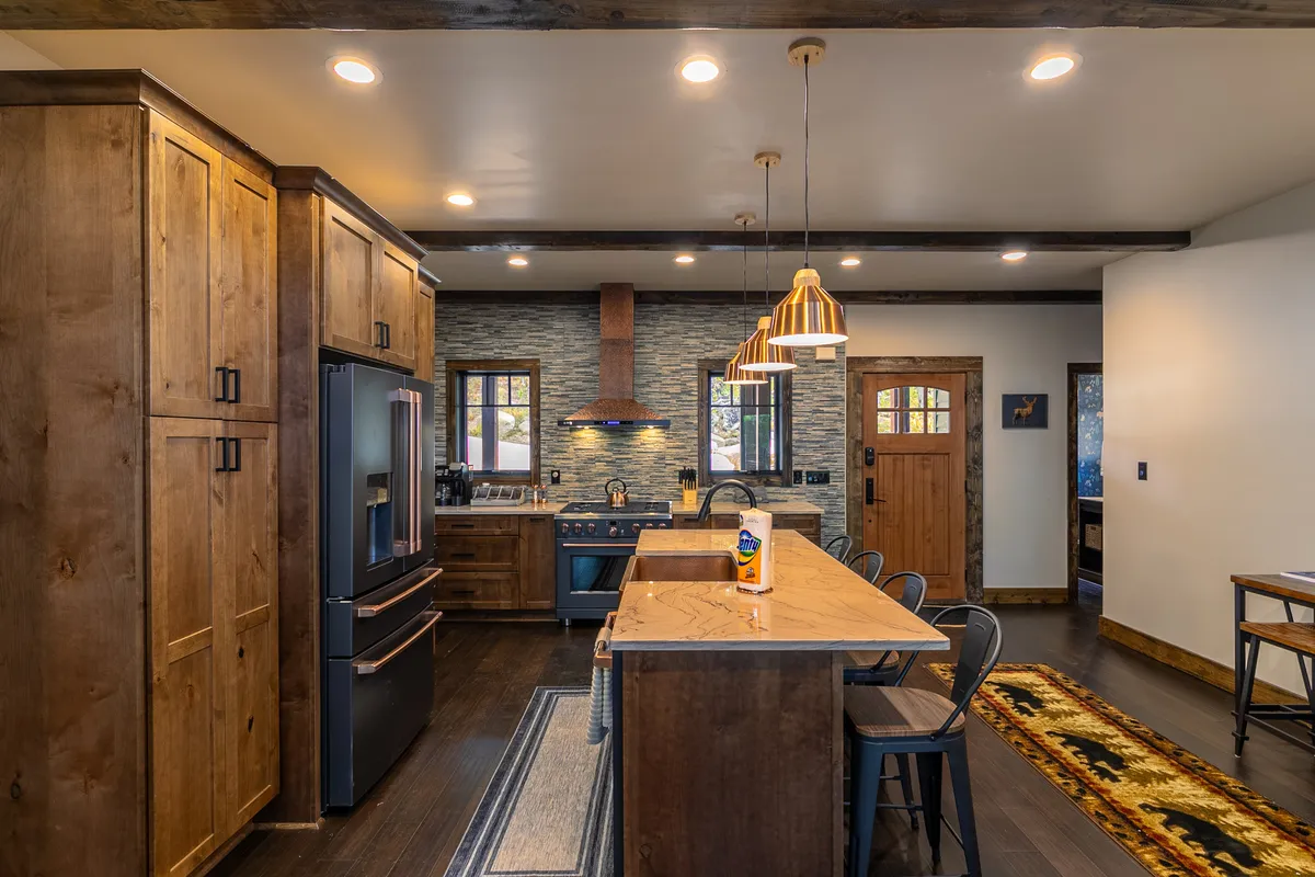 This image showcases a warm, rustic kitchen with wooden cabinets, a large kitchen island, stainless steel appliances, pendant lights, and a stone backsplash. The space looks spacious, inviting, and clearly designed for group meal prep and gatherings.