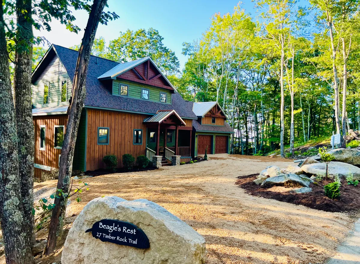 This image captures the house facade surrounded by tall trees, with a prominent stone sign and a peaceful yard. The setting suggests tranquility and connection with nature, inviting guests to relax or explore nearby trails.