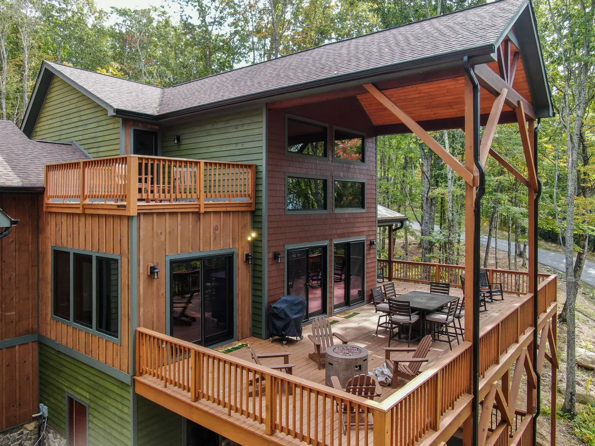 A wide, welcoming wooden deck attached to the home, with outdoor dining furniture set up for group gatherings. Large windows and natural wood finishes add warmth, and the deck is surrounded by forested greenery, suggesting space for both relaxation and socializing.