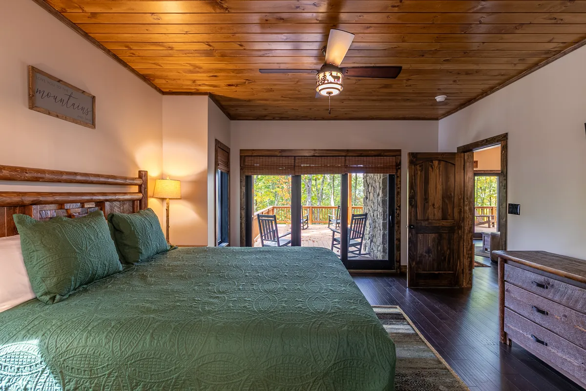 A serene primary bedroom suite featuring a king-size bed with a soothing green quilt, wooden ceiling, and expansive windows that frame tranquil outdoor views. The space exudes quiet luxury and comfort, with an inviting atmosphere ideal for unwinding after a day of adventure.