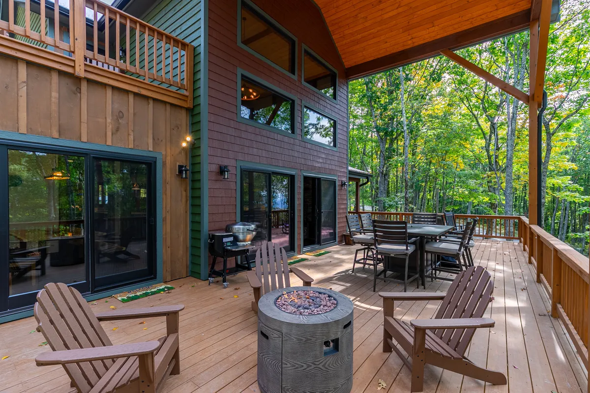 This exterior image showcases a spacious deck with comfortable seating, a fire pit, and large windows looking into the house. The area is surrounded by trees, creating a secluded and inviting outdoor living space.