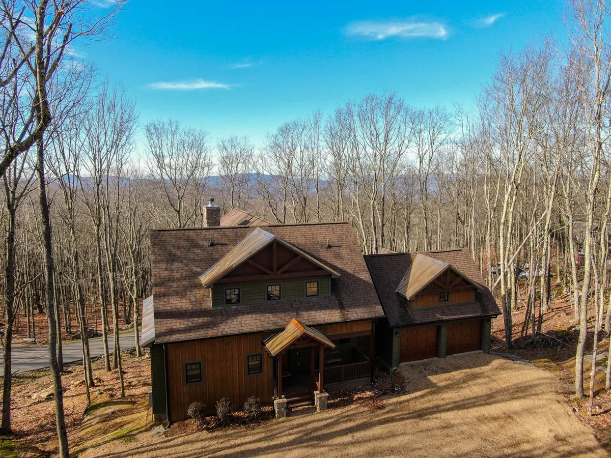 The exterior of a luxurious wooden home nestled among trees, featuring a sloped roof and distant mountain vistas. The setting suggests peace, seclusion, and a close connection with the surrounding natural beauty, as well as ample space for outdoor gatherings or quiet reflection.