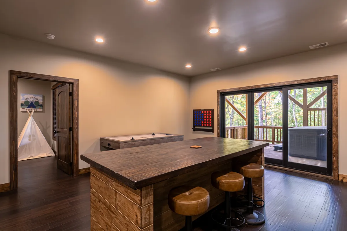 A lively interior scene featuring a bar counter, an air hockey table, and large windows that open to a deck. The space radiates energy and fun, with game tables set up for play and a backdrop of natural light streaming in. Colorful game boards and social seating make this room ideal for group entertainment.
